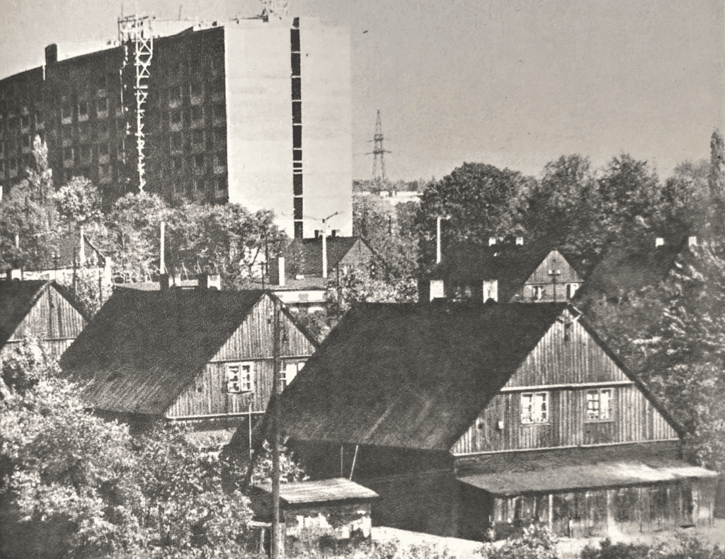 Czerno- białe zdjęcie przedstawiające szereg domków kunitzerowskich. W tle widać wieżowiec