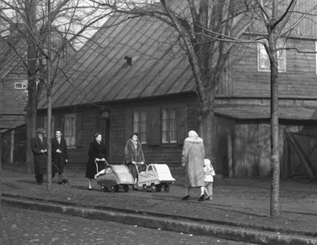 Czerno- białe zdjęcie przedstawiające szereg domków kunitzerowskich. Po ulicy spacerują przechodnie. 