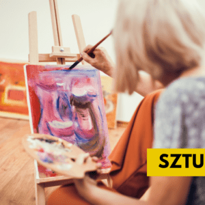 Starsza kobieta maluje obraz farbami. Płótno stoi na drewnianej sztaludze. W tle widać inne obrazy.