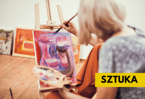 Starsza kobieta maluje obraz farbami. Płótno stoi na drewnianej sztaludze. W tle widać inne obrazy.