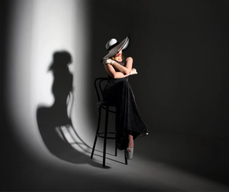 Kobieta w czerni pozująca do zdjęcia. Na głowie ma kapelusz. W tle jej cień.